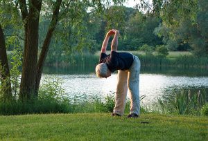 Ein älterer Mann bei Dehnungsübungen auf einer Wiese vor einem See, Stichwort Beweglichkeit. (c) Pixabay.com