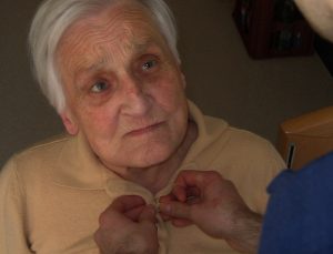 Das Gesicht einer alten Frau, der von einer 24-Stunden-Betreuung die Weste zugeknöpft wird. (c) Pixabay.com