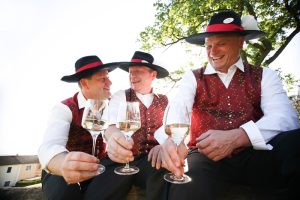 Mostwiki.at – altes Wissen über die Region Moststraße 1 Drei Männer in Tracht mit einem Glas Wein, Stichwort MostWiki. (c) Schwarz-König.at
