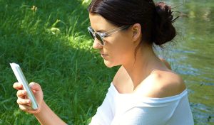 Eine Frau mit Sonnenbrille am Wasser auf ihr Handy schauend, Stichwort Apps am Handy. (c) Pixabay.com