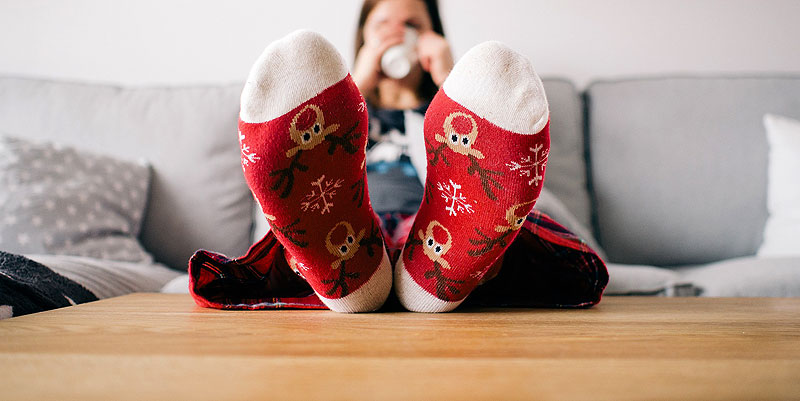 Eine Frau mit Socken mit Weihnachtsmuster sitzt, die Füße am Tisch mit einer Tasse Tee gemütlich auf der Couch. (c) Pixabay.com