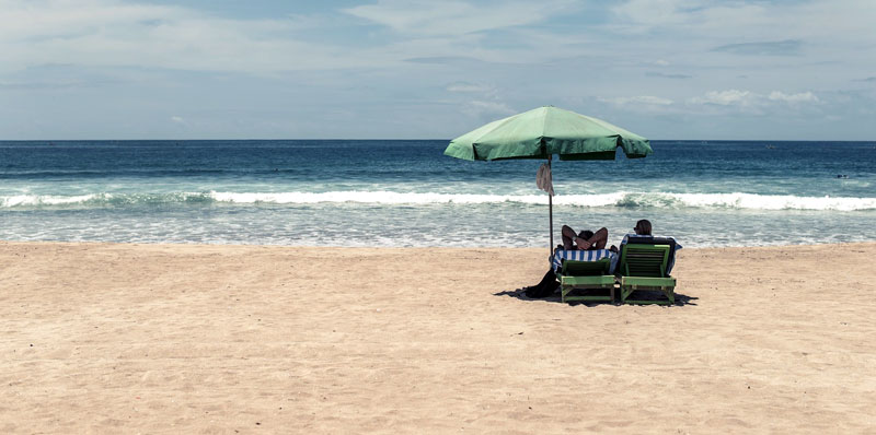 Zwei Menschen auf Liegen unter einem Sonnenschirm auf einem einsamen Sandstrand. (c) Pixabay.com