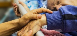 Eine alte zu pflegende Person in einem Krankenbett mit einer Hand am Rausfallschutz, die von einer anderen Person gehalten wird. (c) Pixabay.com