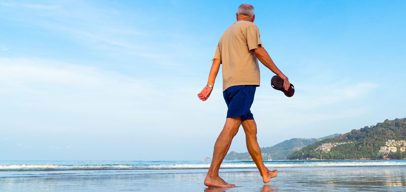 Ein älterer Mann geht barfuß über einen Strand und schaut auf´s Meer. (c) Pixabay.com