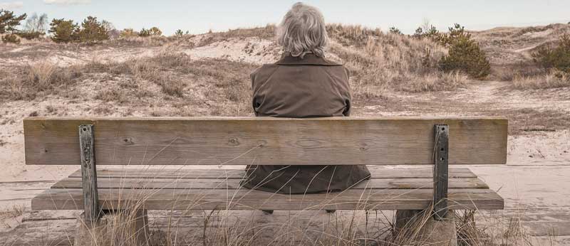 Eine ältere Person von hinten sitzt auf einer Bank und schaut in die karge Landschaft. (c) Pixabay.com
