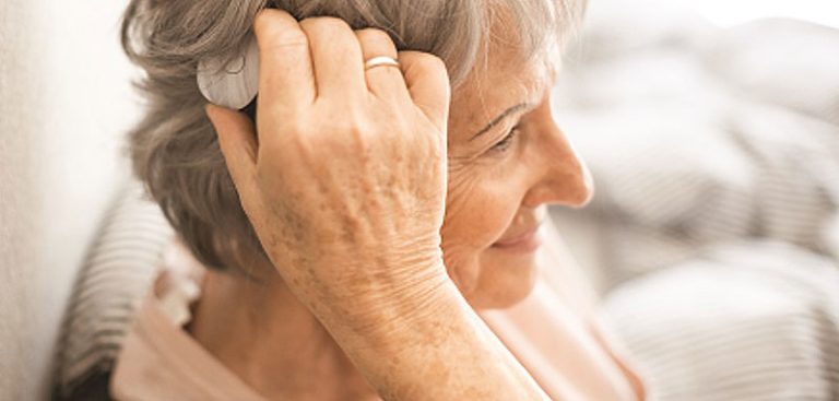 Eine alte Frau greift mit der Hand zum Audioprozessor ihres Cochlea-Implantats. (c) MED-EL
