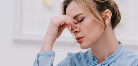 Eine von Kopfschmerzen geplagte Frau greift sich mit der Hand auf den Nasenrücken. (c) Protina Pharmazeutische GmbH/ IgorVetushko