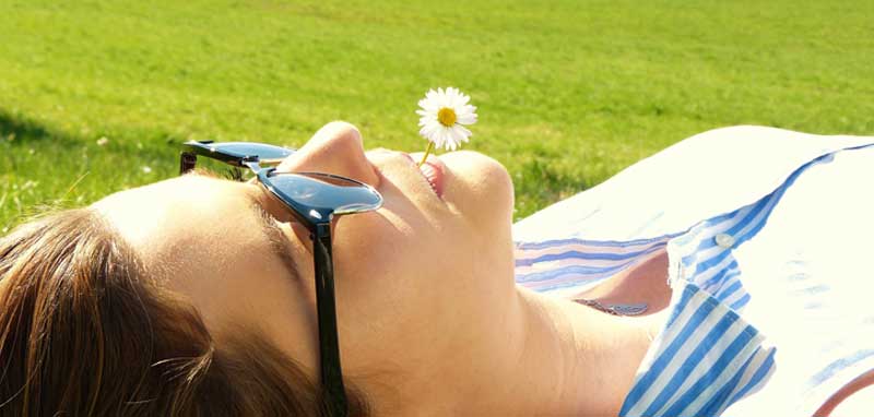 Eine Frau mit Sonnenbrille und Gänseblümchen im Mund liegt auf einer Wiese. (c) Pixabay.com