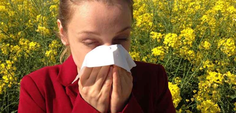Eine Frau mit Heuschnupfen putzt sich vor einer blühenden Wiese stehend die Nase. (c) Pixabay.com