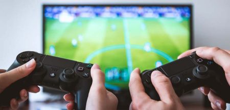 Die Hände einer Frau und eines Mannes mit Kontroller einer Spielkonsole, dahinter auf einem Fernseher ein Fußballspiel. (c) Pixabay.com