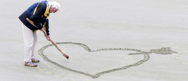 Ein alter Mann, der mit einem Stock ein Herz in einen Sandstrand zeichnet. (c) Pixabay.com
