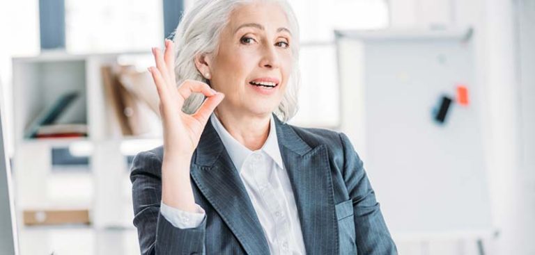Eine ältere Frau mit grauen Haaren sitzt in einem Büro und lacht in die Kamera. (c) Protina Pharmazeutische GmbH.