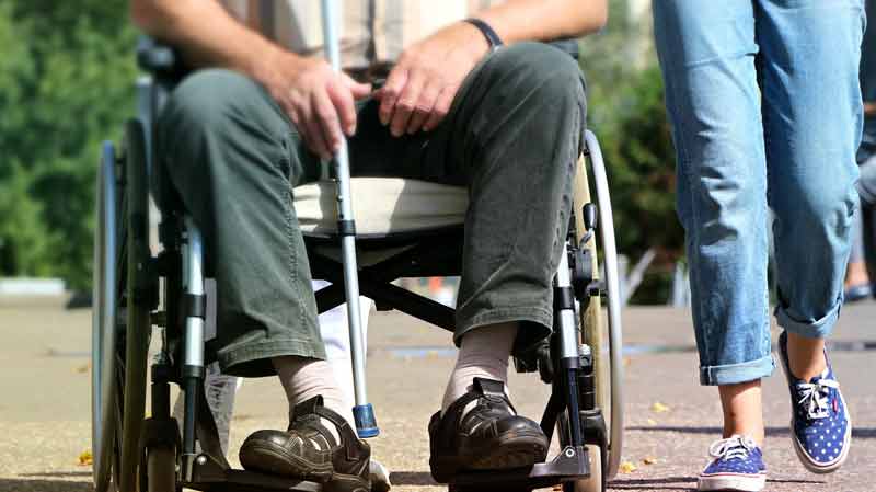 Die Füße eines Mannes in einem Rollstuhl, daneben geht eine Frau, Stichwort pflegende Angehörige. c) Pixabay.com