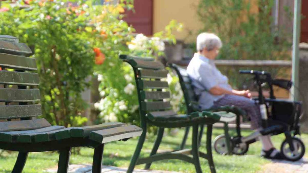 Eine alte Frau sitzt auf einer Parkbank, neben ihr steht ihr Rollator. (c) Pixabay.com