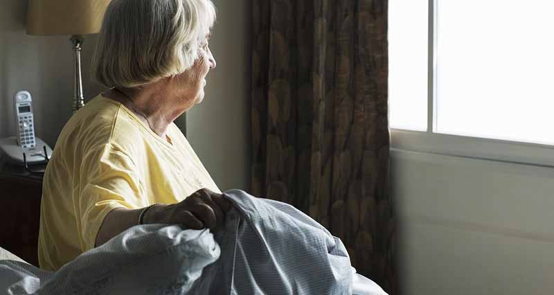 Eine alte Frau sitzt auf ihrem Bett und schaut beim Fenster raus. (c) Pixabay.com