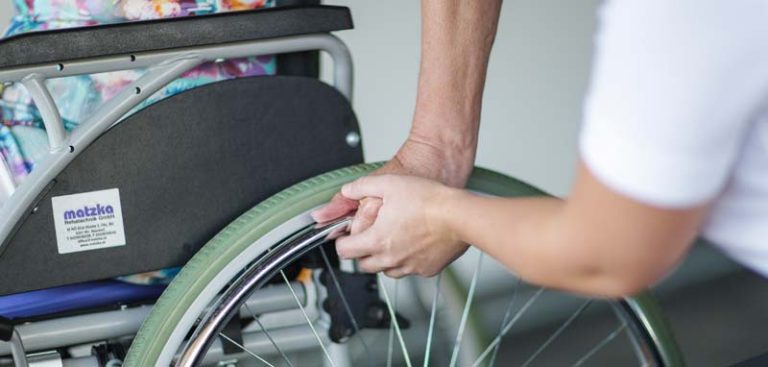 Ergotherapie: eine Therapeutin hält die Hand einer Patientin, die im Rollstuhl sitzt, am Rad, um sie beim Anschieben zu unterstützen. (c) Schultz & Schultz/ Ergotherapie Austria