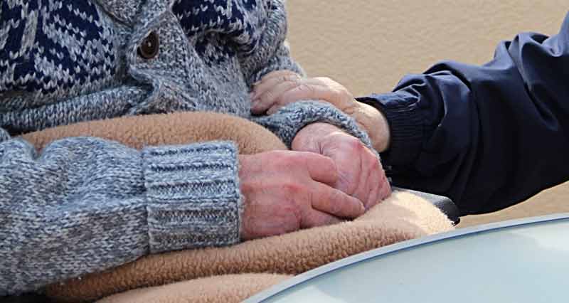Die Arme einer alten sitzenden Person, die mit einer Decke zugedeckt ist. Die Hand einer anderen Person hält tröstend den Unterarm. (c) Pixabay.com