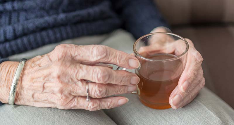 Die Hände einer alten Frau, die ein Glas mit Tee auf ihren Beinen hält. (c) Pixabay.com