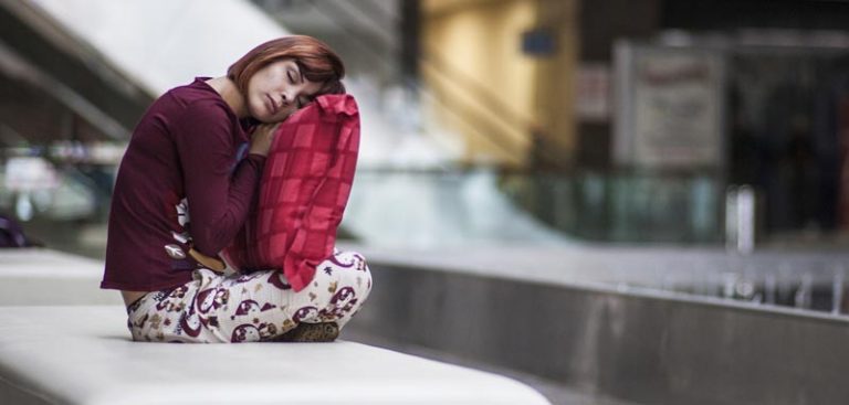 Eine Frau in Pyjama sitzt in einem öffentlichen Gebäude auf einer Bank und hat ihren Kopf auf einem großen Polster vor ihr. (c) Pixabay.com