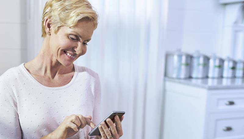 Schwerhörigkeit ist mittlerweile besser behandelbar als je zuvor 1 Eine ältere Frau mit einem Cochlear Implantat – Stichwort Schwerhörigkeit – tippt lächelnd in ihr Smartphone. (c) Cochlear Ltd.
