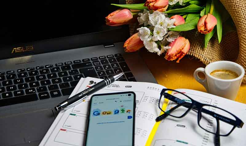 Ein Laptop, davor ein offener Kalender, auf dem ein Kugelschreiber, eine Brille und ein Smartphone liegen; daneben ein Espresso und ein kleiner Blumenstrauß.
(c) Pixabay.com