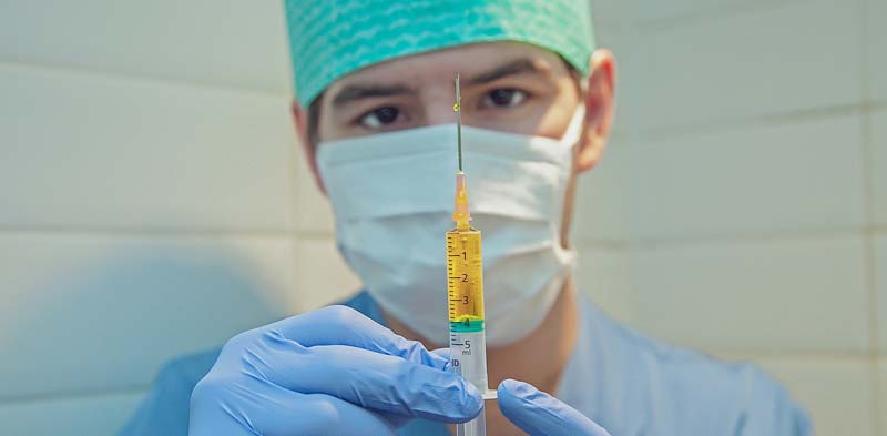 Der Kopf eines Mediziners mit Mundschutz, der eine Spritze vor sich hält, Stichwort Coronavirus-Bekämpfung.
(c) Pixabay.com