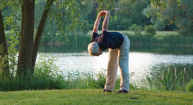 Ein älterer Mann, der auf einer Wiese vor einem See seinen Rücken dehnt, Stichwort Fitness.
(c) Pixabay.com