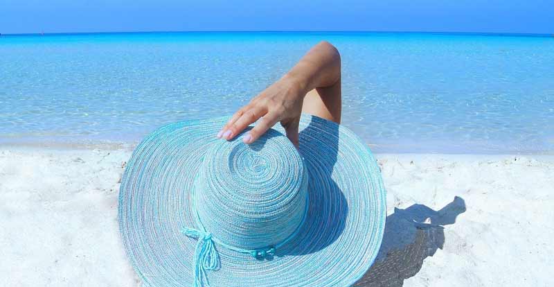 Eine Frau, die mit einer Hand ihren blauen Strohhut hält, auf einem Sandstrand mit Blick auf´s Meer.
(c) Pixabay.com