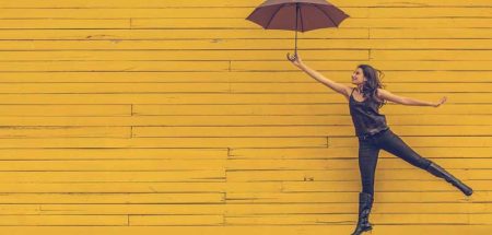 Eine Frau mit Regenschirm macht einen Luftsprung vor einer gelben Wand. (c) Pixabay.com