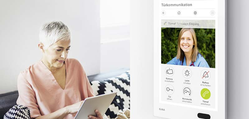 Sehen, wer an der Tür klingelt – eine Seniorin mit einem Tablet auf der Couch beim Öffnen der Haustür mittels Türkommunikationssystem inkl. Kamera. (c) www.gira.at
