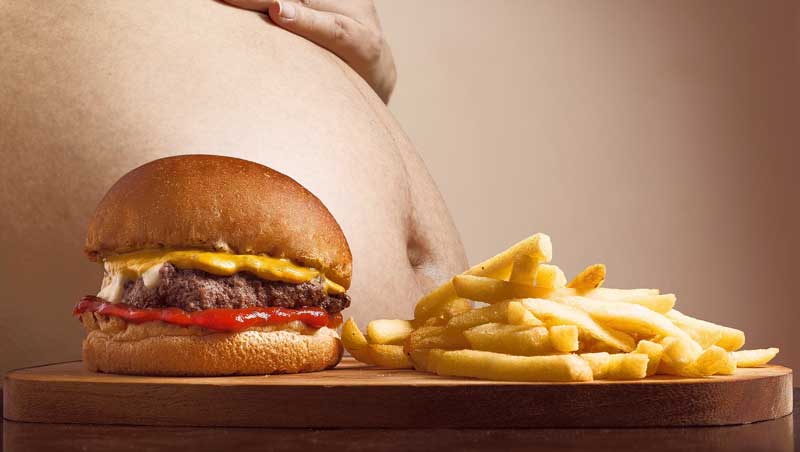 Ein großer Burger neben fetten Pommes, dahinter der dicke Bauch eines Mannes.
(c) Pixabay.com