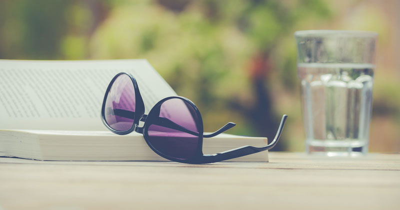 Eine Sonnenbrille auf einem aufgeschlagenen Buch, dahinter ein Wasserglas, Stichwort reisen.
(c) Pixabay.com