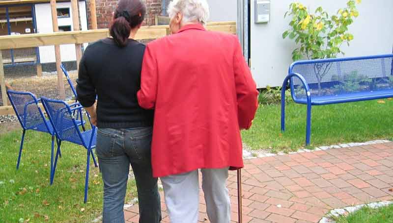 Eine alte Frau mit Gehstock geht eingehängt im Arm einer anderen Frau auf einem Gehweg.
(c) Pixabay.com
