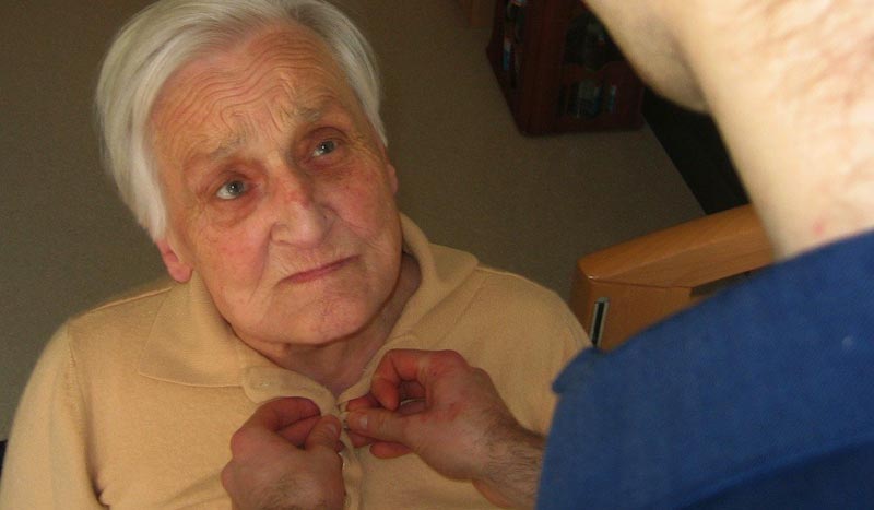 Das Gesicht einer alten Frau, der ein Pfleger ihre Bluse zuknöpft.
(c) Pixabay.com