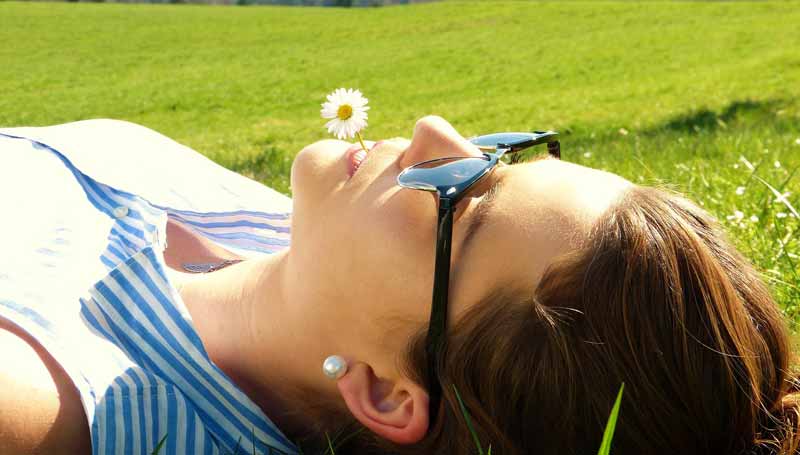 Eine Frau mit Sonnenbrille und einem Gänseblümchen im Mund liegt auf einer Wiese.
(c) Pixabay.com