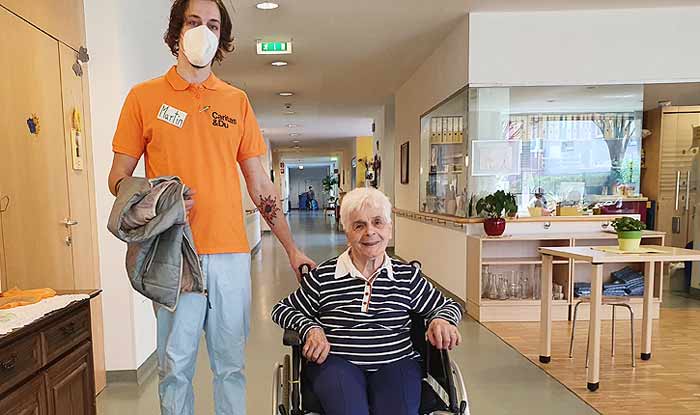 Ein Pfleger steht neben einer alten Frau im Rollstuhl in einem Altenheim.
(c) Caritas