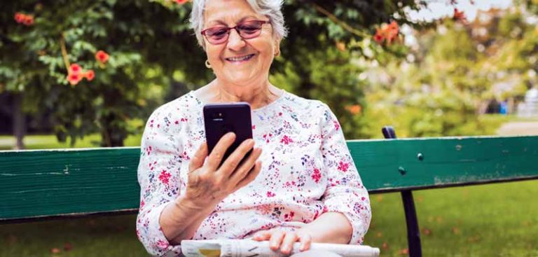 Eine ältere Frau lächelnd auf einer Parkbank auf ihr Smartphone schauend. (c) tarife.at