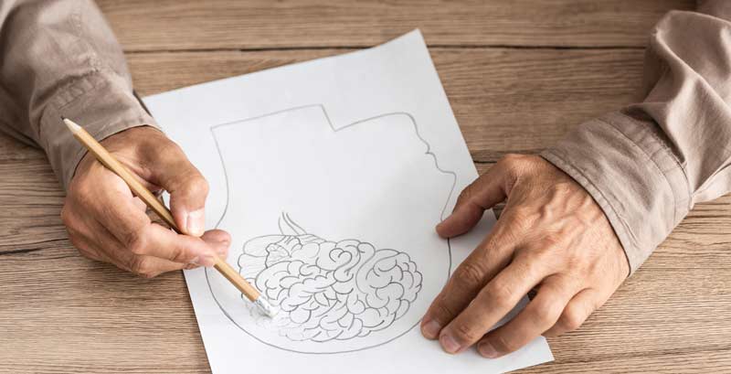 Die Hände eines Mannes, der das Gehirn auf einer Zeichnung vor sich wegradiert, Stichwort Alzheimer-Symptome.
(c) AdobeStock