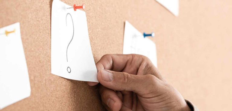 Die Hand eines Mannes, der einen Zettel mit einem Fragezeichen auf einer Pinwand hält. (c) AdobeStock