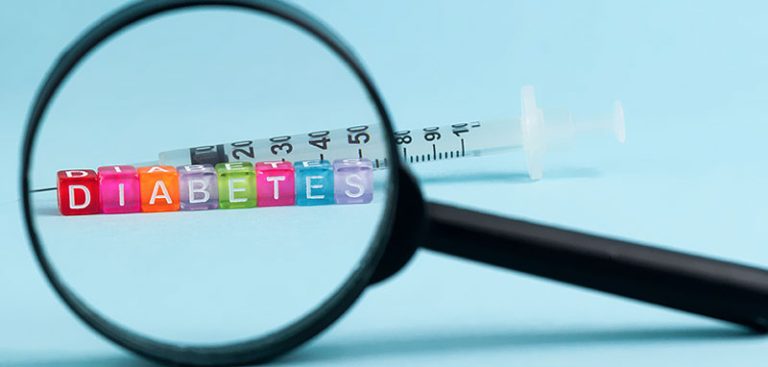 Das Wort Diabetes mit bunten Scrabble-Würfeln, dahiner eine Spritze – beides durch eine Lupe gesehen. (c) AdobeStock