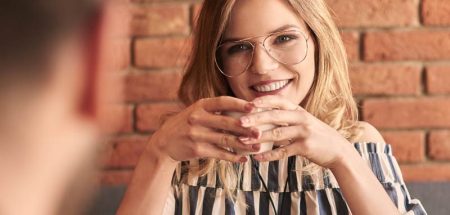Eine lächelnde Frau mit Brille mit einem Glas in den Händen an einem Tisch. (c) Seeking.com