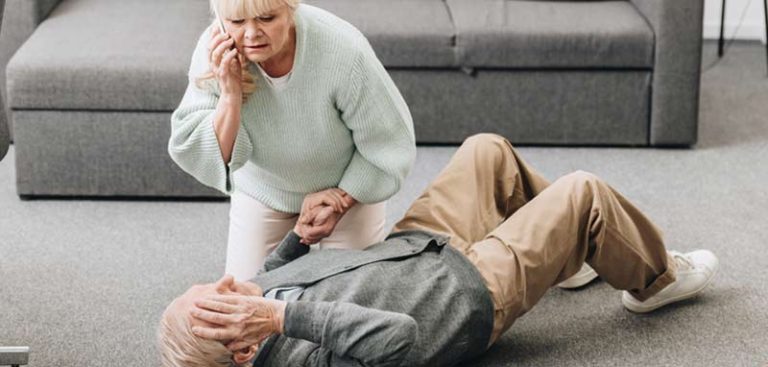 Ein alter Mann liegt auf dem Teppich, daneben kniet seine Frau, die seine Hand hält und Hilfe ruft. (c) AdobeStock
