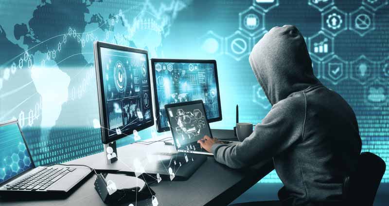 Ein Mann mit Kapuze über dem Kopf vor einem Laptop und zwei großen Monitoren, Stichwort Cyberschutz.
(c) AdobeStock
