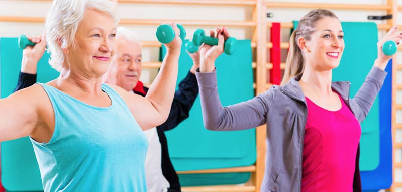 Zwei Senior•innen mit einer jüngeren Frau, alle auf einem Gymnastikball, machen Übungen mit Kurzhanteln. (c) AdobeStock