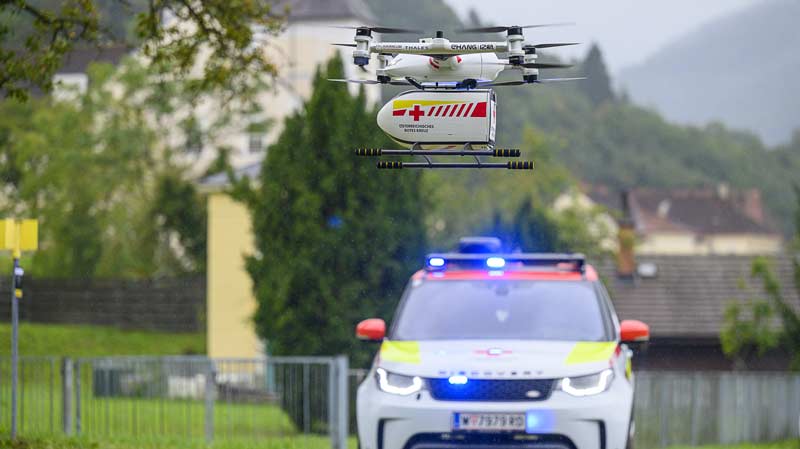 Eine fliegende Drohne mit einer Blutkonserve, dahinter ein Wagen vom Roten Kreuz.
(c) ÖRK/ pixelmaker.at