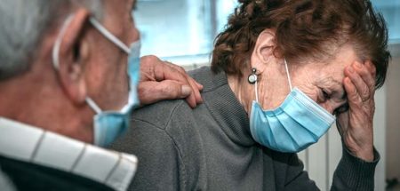 Ein alter Mann, der einer alten Frau die Hand auf ihre Schulter legt; beide mit Mund-Nasen-Schutz. (c) AdobeStock