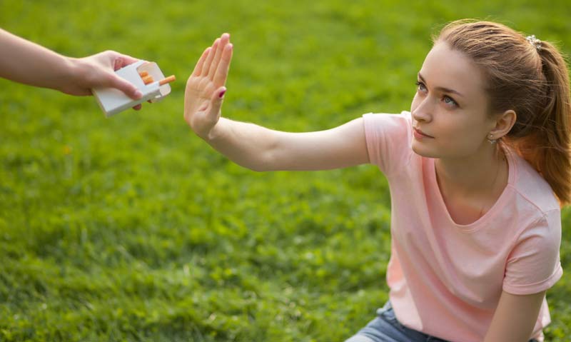 Eine junge Frau sitzt im Gras und macht mit ihrer Hand ein Stop-Zeichen zu jemanden, der ihr eine Zigarette anbietet.
(c) AdobeStock