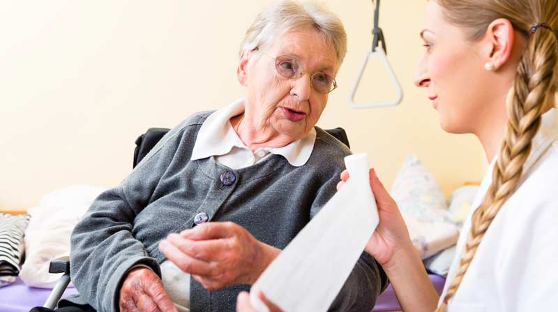 Eine alte Frau neben einer Pflegerin, die ihr einen neuen Verband anlegen möchte, Stichwort Wundbehandlung.
(c) AdobeStock