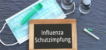 Ein Tafel mit "Influenza Schutzimpfung", daneben ein Mund-Nasen-Schutz, eine Spritze und ein Serum. (c) AdobeStock