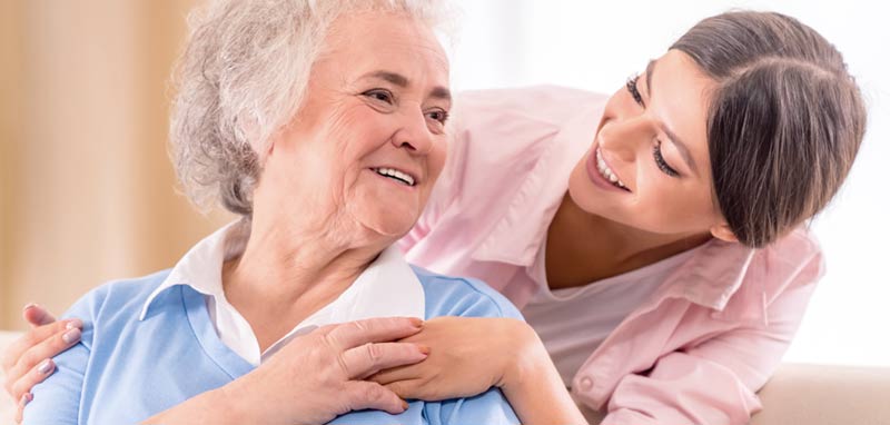 Eine Betreuerin, die einer älteren Frau von hinten über die Schulter greift und mit ihr spricht. (c) AdobeStock